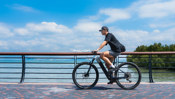 A cyclist rides on a coastal road with blue sky, clouds, and water in the background.